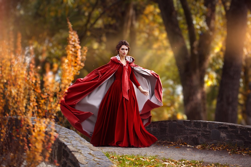 Czerwona suknia ślubna. Kobieta w czerwonej sukni i pelerynie idzie po parku.