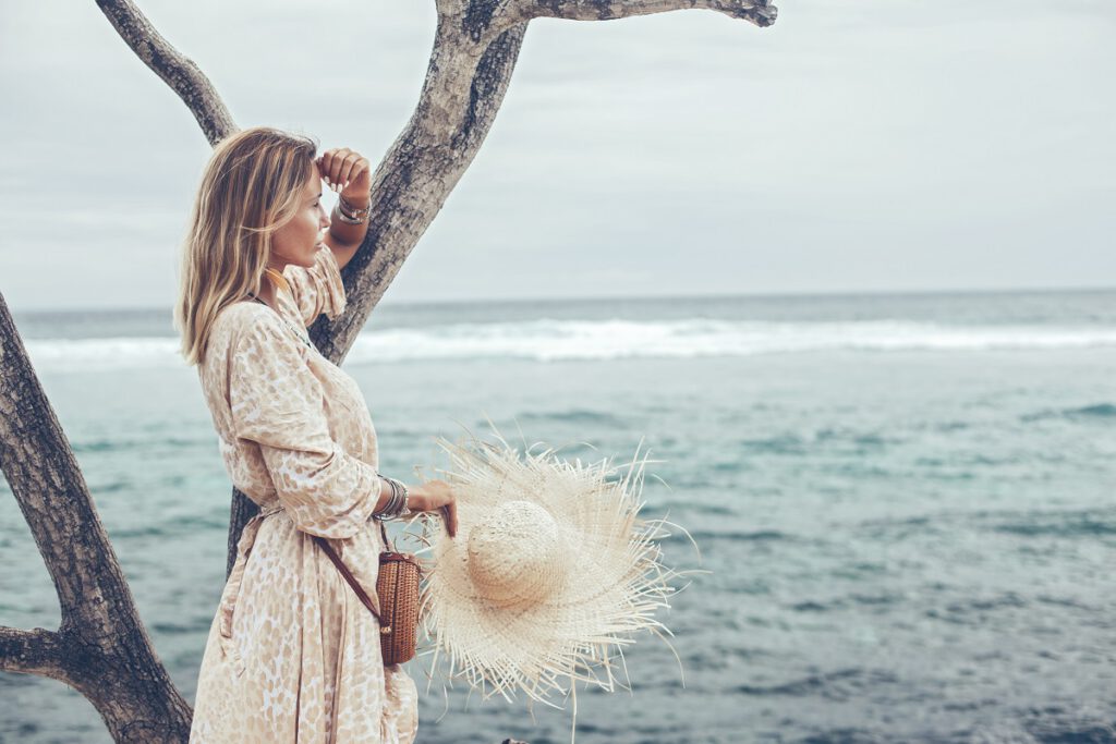 Sukienki boho na wesele - blondynka z kapeluszem patrzy na morze
