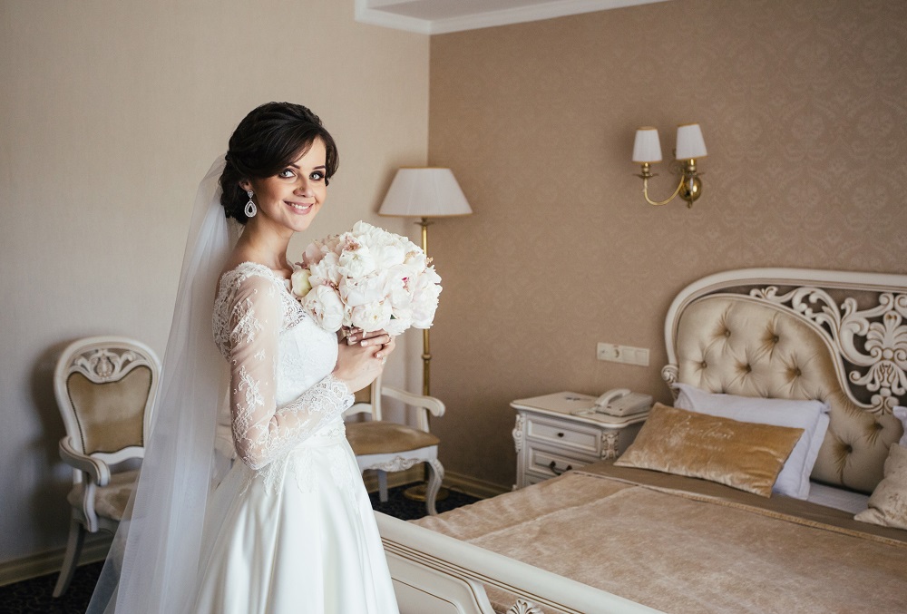 suknia ślubna z długim rękawem - panna młoda pozuje do zdjęcia z bukietem w sypialni