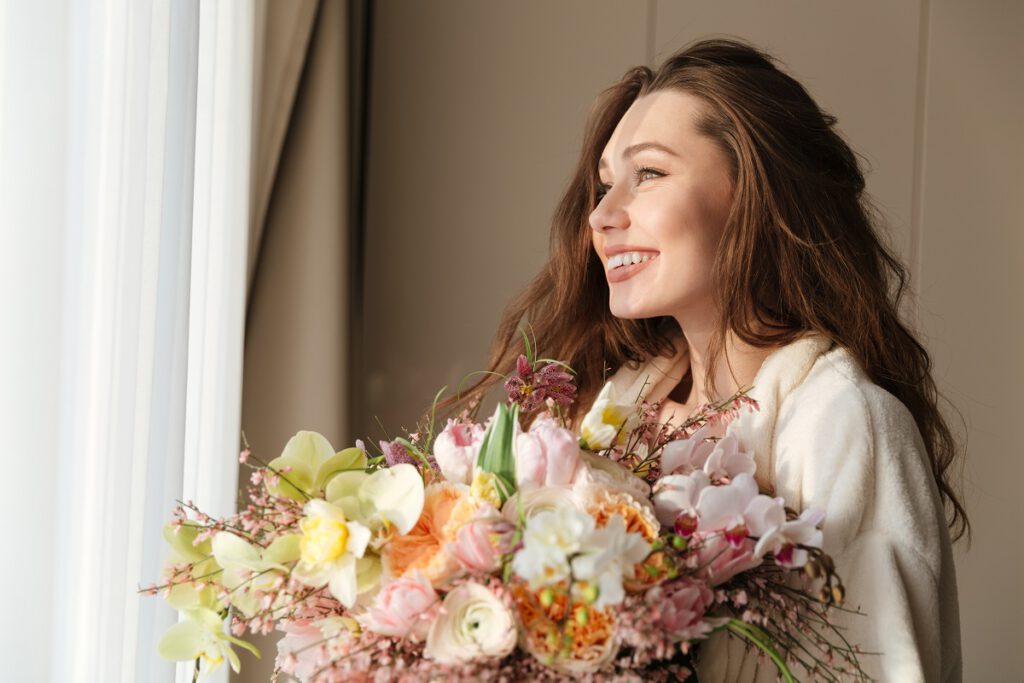 prezent na dzień matki-uśmiechnięta kobieta stoi i patrzy przez okno trzymając bukiet kwiatów